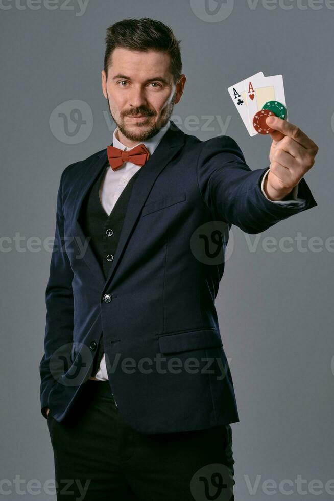 homem dentro Preto clássico terno e vermelho gravata-borboleta mostrando dois jogando cartões e salgadinhos, posando em cinzento estúdio fundo. jogatina, pôquer, casino. fechar-se. foto