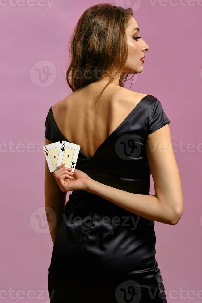 morena fêmea dentro Preto vestir. mostrando dois jogando cartões, posando em pé costas para Câmera contra Rosa fundo. pôquer, casino. fechar-se foto
