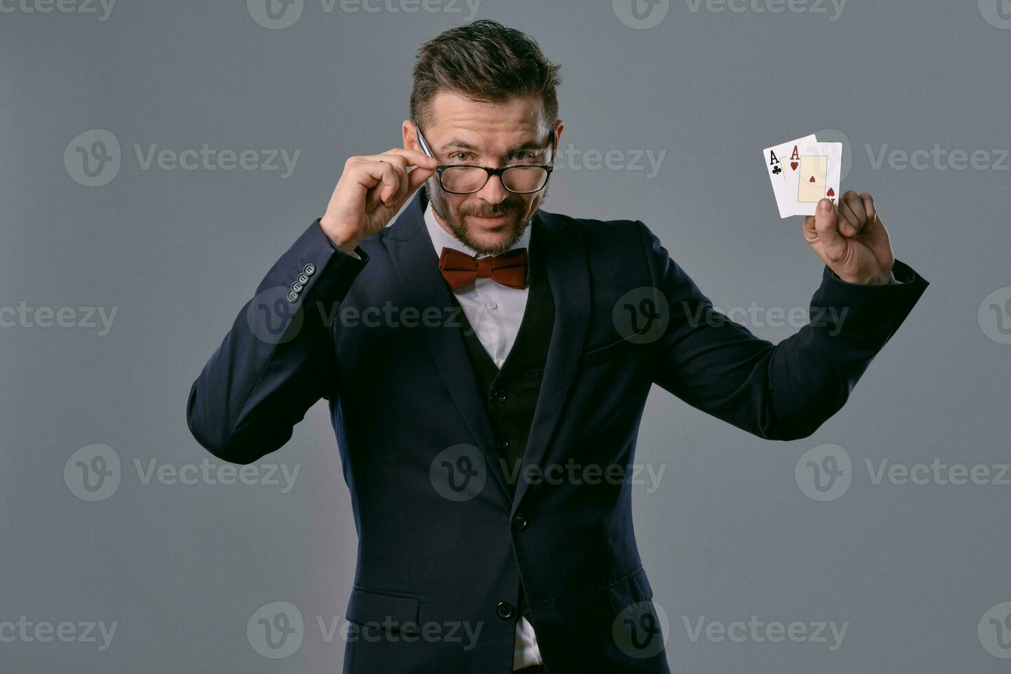 homem dentro Preto clássico terno, vermelho gravata borboleta, glases é mostrando dois jogando cartões, posando em cinzento estúdio fundo. jogatina, pôquer, casino. fechar-se. foto