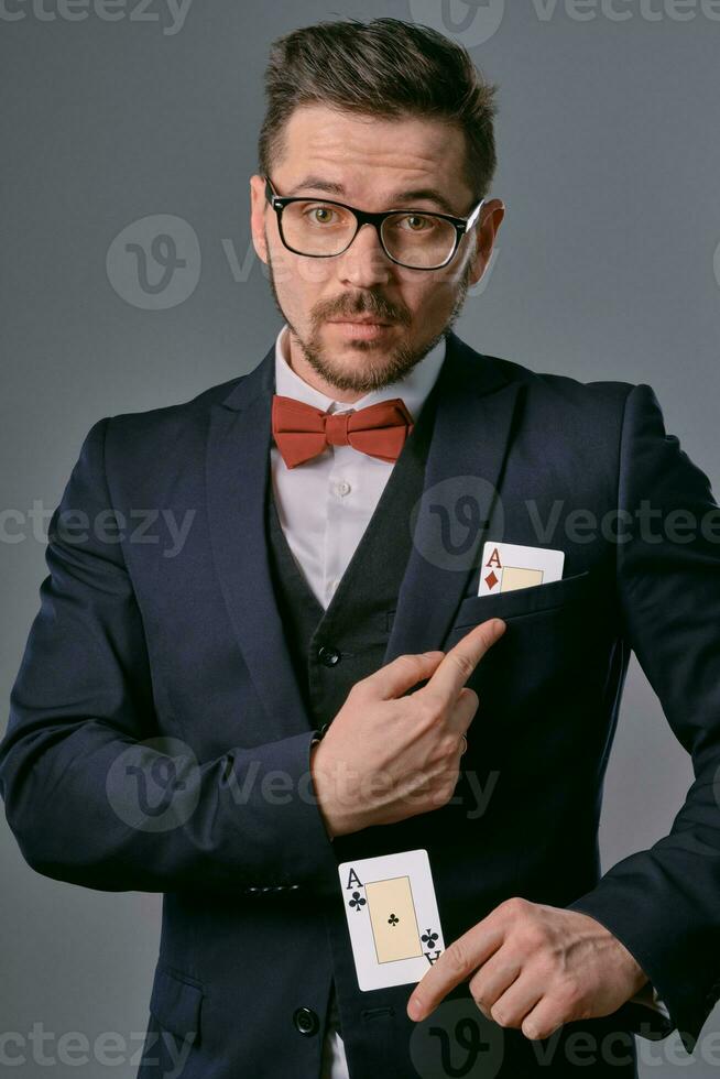 homem dentro Preto clássico terno, vermelho gravata borboleta, glases é mostrando dois jogando cartões, posando em cinzento estúdio fundo. jogatina, pôquer, casino. fechar-se. foto