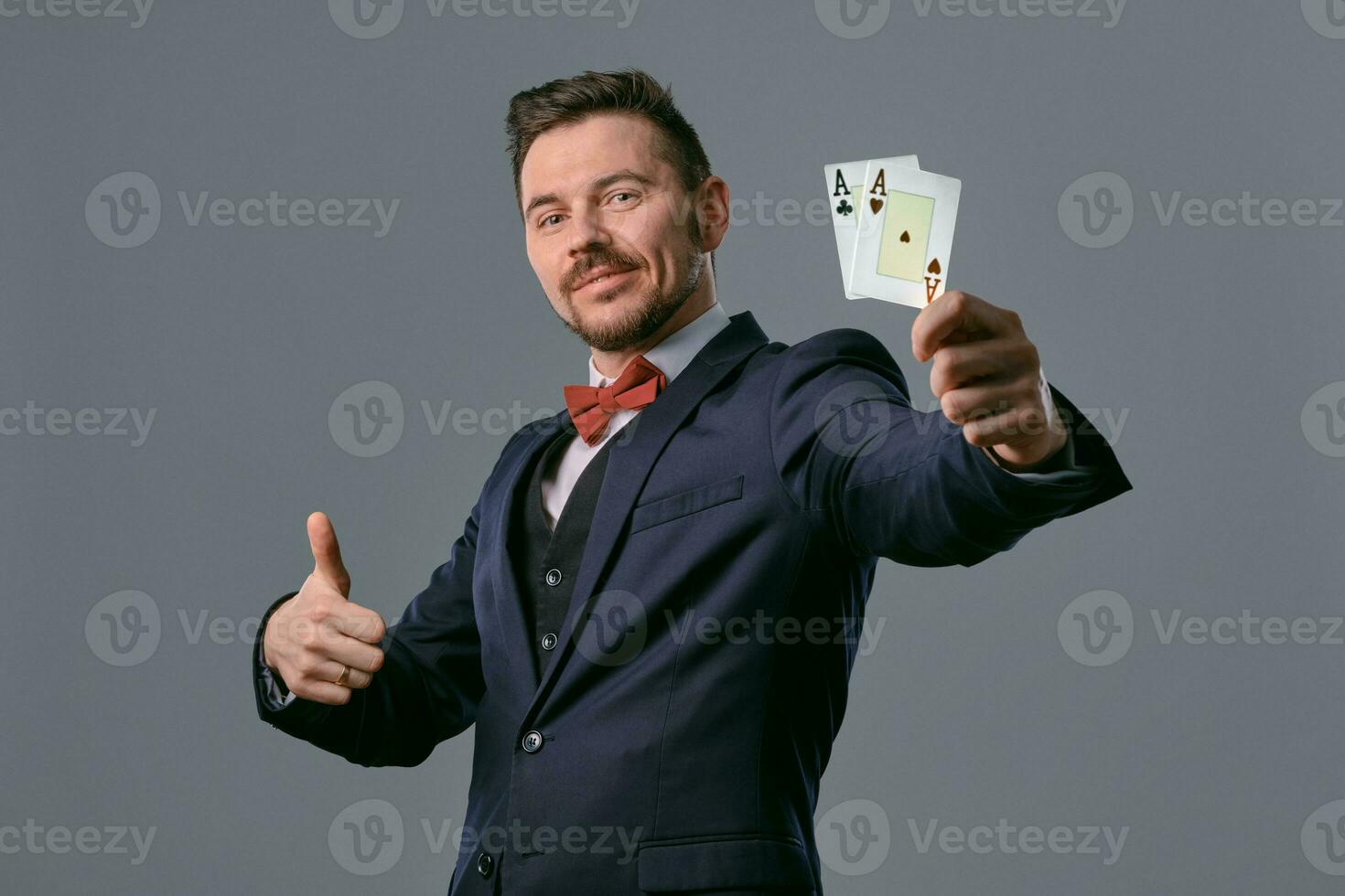 homem dentro Preto clássico terno e vermelho gravata-borboleta mostrando dois jogando cartões enquanto posando contra cinzento estúdio fundo. jogatina, pôquer, casino. fechar-se. foto