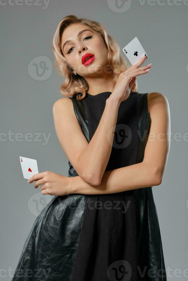 Loiras menina dentro Preto couro vestir mostrando dois jogando cartões, posando contra cinzento fundo. jogos de azar entretenimento, pôquer, casino. fechar-se. foto