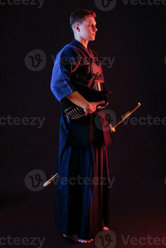 kendo lutador vestindo dentro a armaduras, tradicional quimono é segurando dele capacete e Shinai bambu espada enquanto posando em uma Preto fundo. fechar acima. foto
