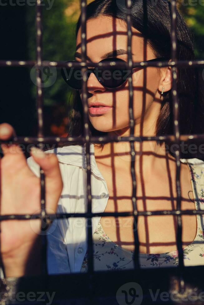 retrato do menina dentro oculos de sol posando dentro cidade atrás uma treliçado cerca. vestido dentro topo com floral imprimir, branco camisa, Preto calça, cintura bolsa. foto