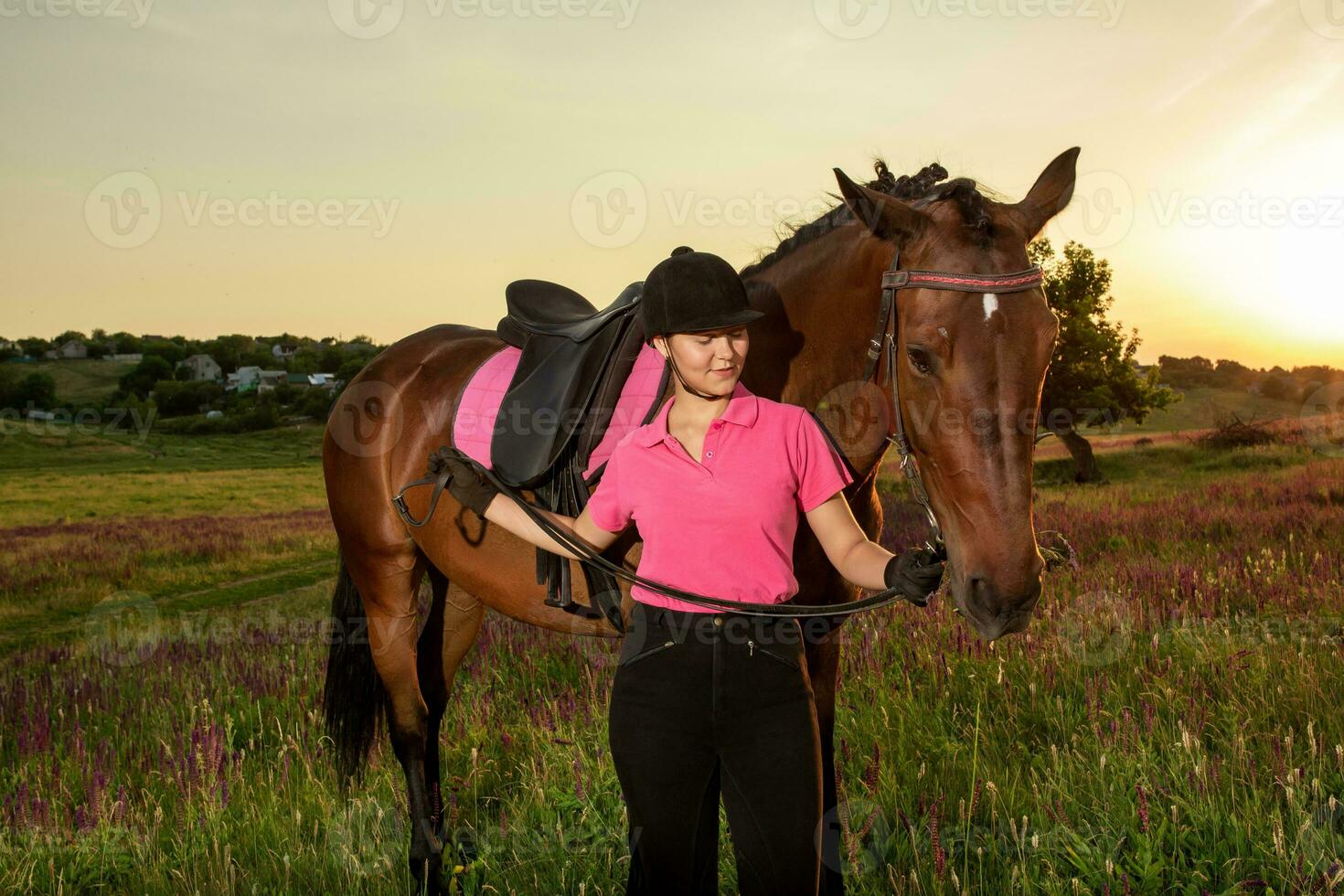 lindo sorridente menina jóquei ficar de pé Próximo para dela Castanho cavalo vestindo especial uniforme em uma céu e verde campo fundo em uma pôr do sol. foto