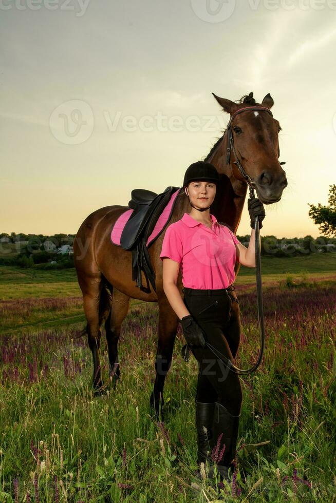lindo sorridente menina jóquei ficar de pé Próximo para dela Castanho cavalo vestindo especial uniforme em uma céu e verde campo fundo em uma pôr do sol. foto