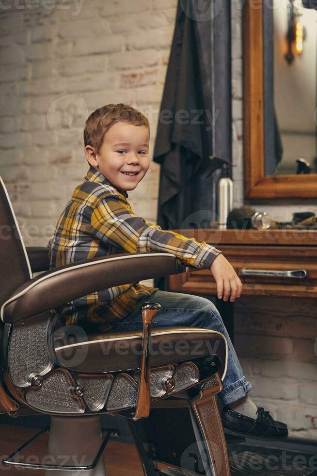 retrato do uma à moda pequeno Garoto vestido dentro camisa e jeans dentro a barbearia, sentado dentro uma cadeira contra a barbearia local de trabalho foto