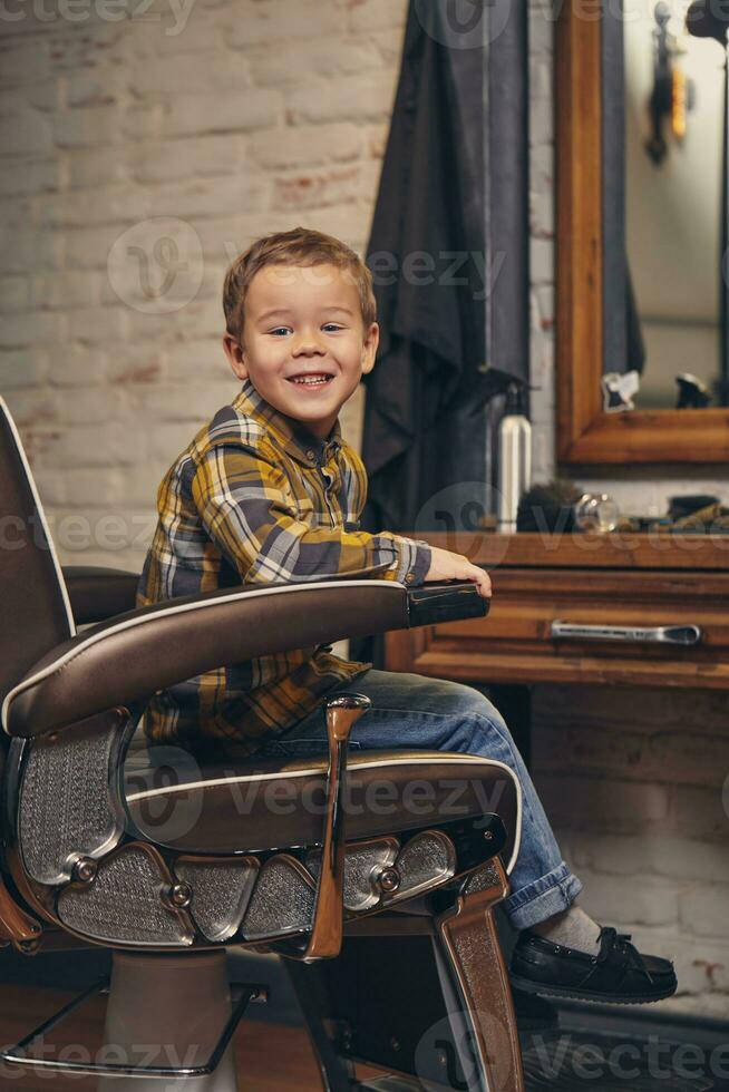 retrato do uma à moda pequeno Garoto vestido dentro camisa e jeans dentro a barbearia, sentado dentro uma cadeira contra a barbearia local de trabalho foto