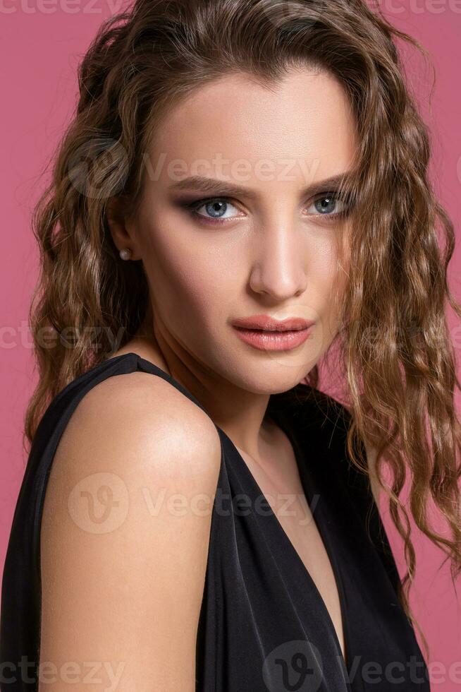 lindo elegante menina com grandes encaracolado cabelo dentro uma Preto vestir dentro a estúdio em uma Rosa fundo. foto