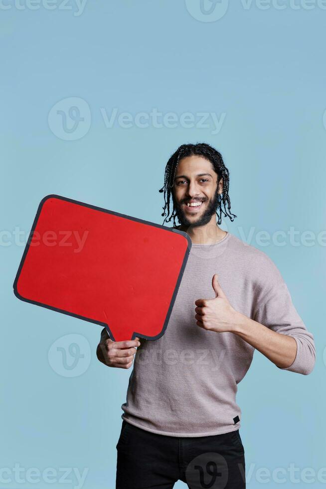 sorridente árabe homem segurando vermelho em branco bandeira com cópia de espaço e mostrando polegar acima retrato. pessoa em pé com esvaziar discurso bolha e positivo gesto enquanto olhando às Câmera foto