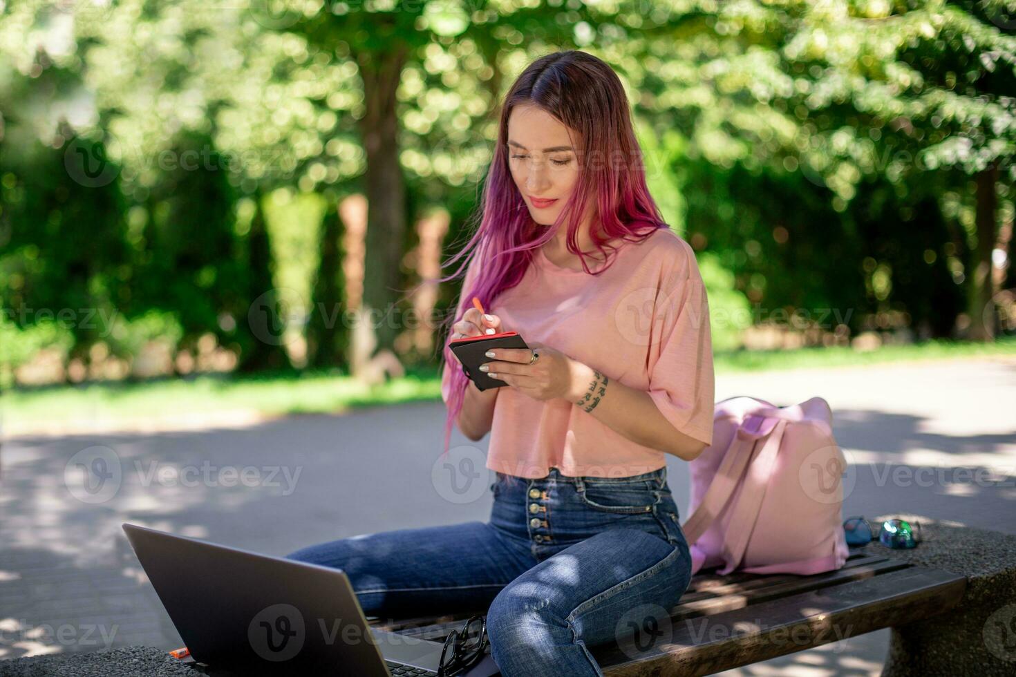 mulher escrevendo dentro uma caderno sentado em uma de madeira Banco dentro a parque. menina trabalhando ao ar livre em portátil computador, cópia de espaço. foto