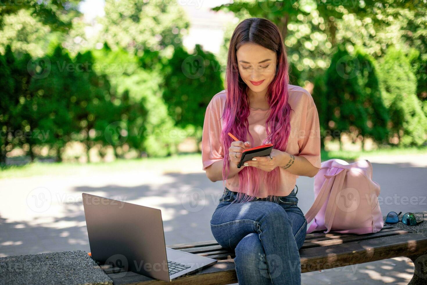 mulher escrevendo dentro uma caderno sentado em uma de madeira Banco dentro a parque. menina trabalhando ao ar livre em portátil computador, cópia de espaço. foto