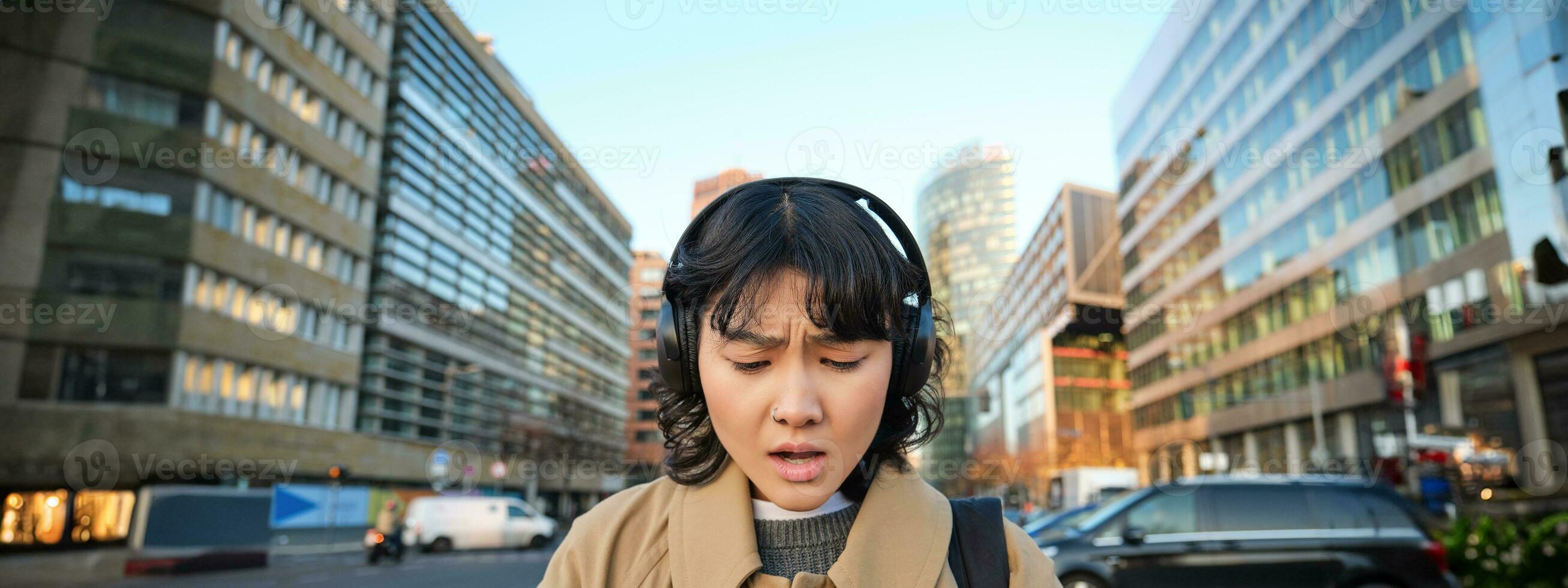 retrato do jovem ásia garota, aluna anda em dentro cidade, escuta música dentro fones de ouvido e usa Móvel telefone em ruas, parece preocupado às tela, lendo mau notícia foto