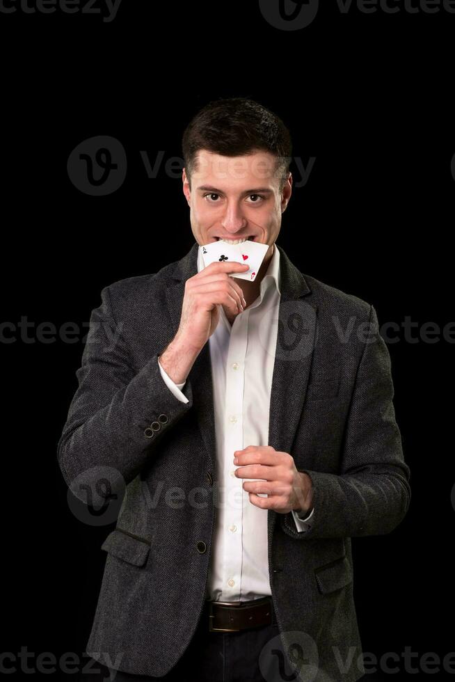 jovem caucasiano homem vestindo terno segurando dois ases dentro dele mão e dentes em Preto fundo. jogos de azar conceito. cassino foto