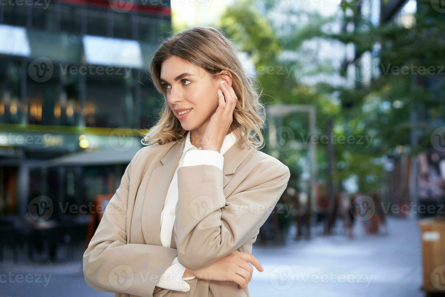 fechar acima retrato do loiro empresária, confiante corporativo mulher dentro fones de ouvido e bege terno, posando ao ar livre em rua do cidade Centro foto