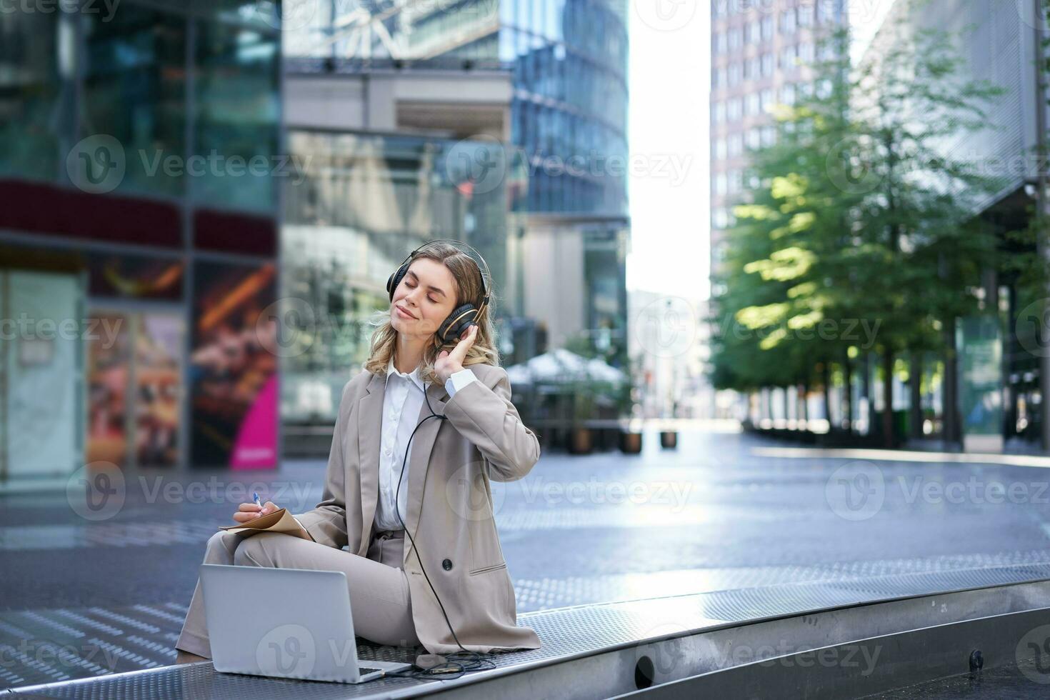 sorridente empresária relaxante, ouvindo para música dentro fones de ouvido a partir de dela computador portátil, sentado sozinho em rua do cidade Centro, vestindo terno foto