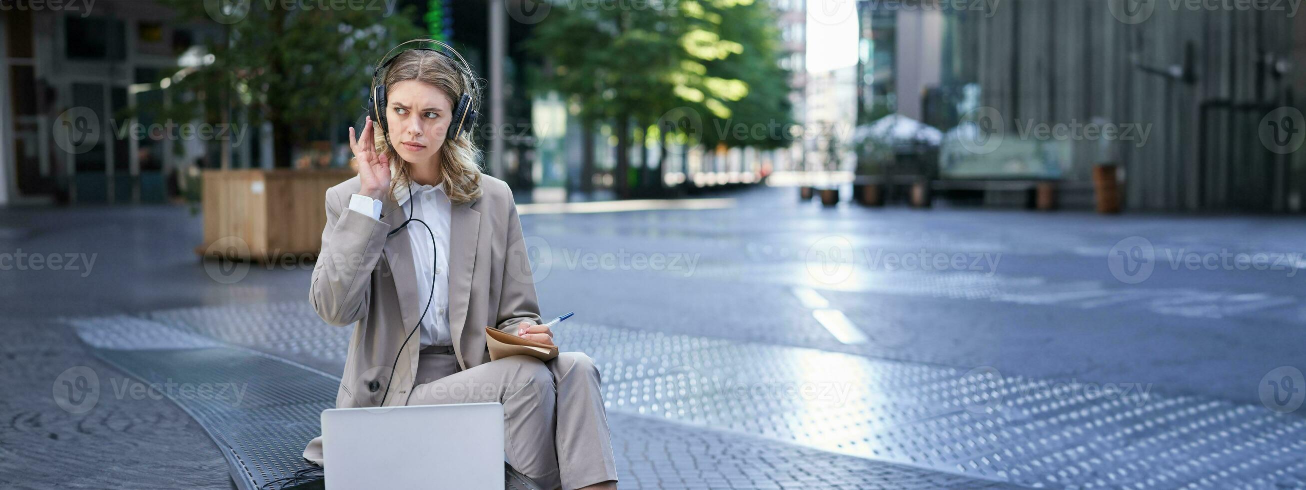 mulher sentado em uma rua com computador portátil e fones de ouvido obstruído em, levando notas. corporativo trabalhador comparecer conectados equipe encontro e escrevendo baixa Informação, trabalhando ao ar livre foto