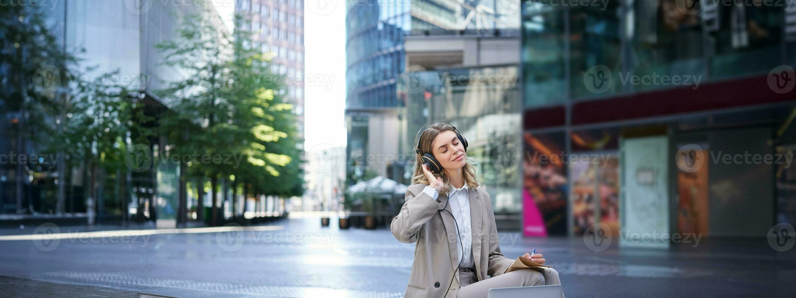 sorridente empresária relaxante, ouvindo para música dentro fones de ouvido a partir de dela computador portátil, sentado sozinho em rua do cidade Centro, vestindo terno foto