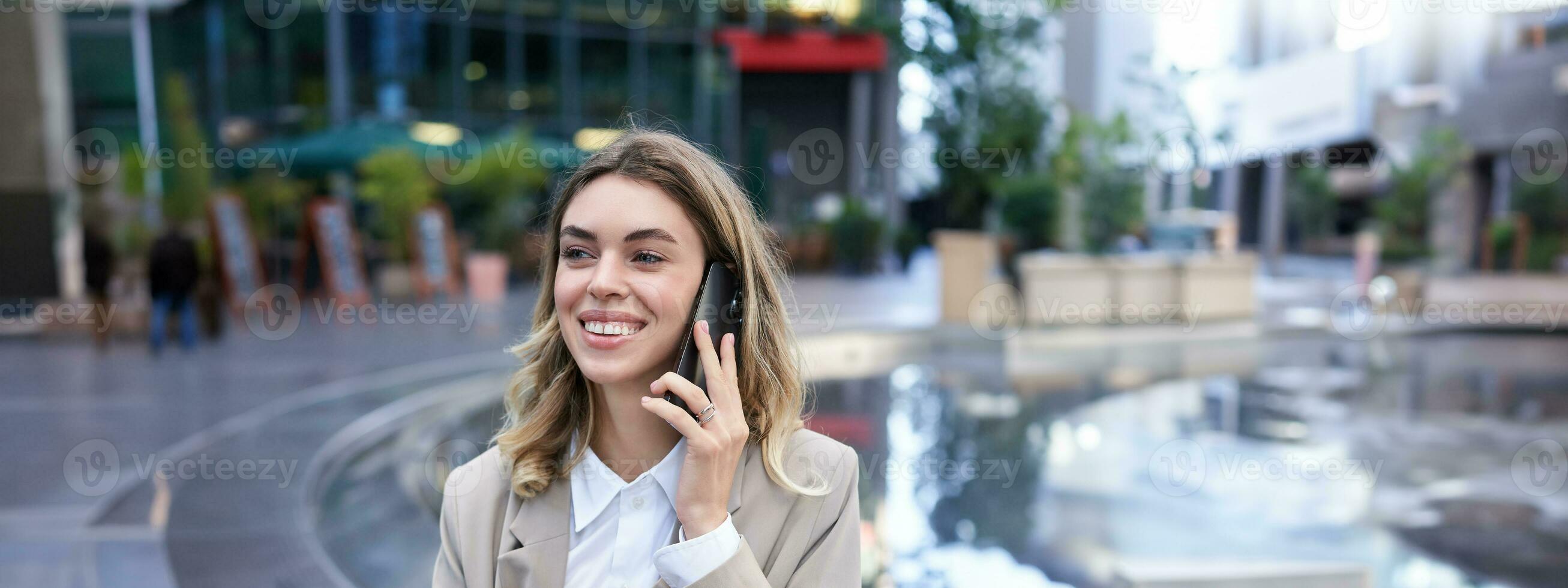 vertical tiro do sorridente mulher falando em Móvel telefone enquanto sentado fora. escritório senhora esperando para alguém depois de trabalhos foto