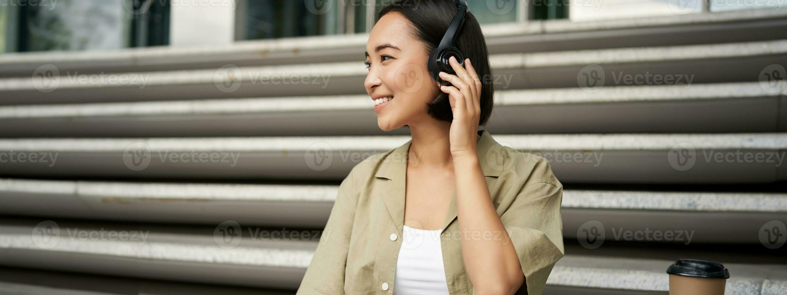 fechar acima retrato do sorridente ásia mulher escuta música dentro fones de ouvido, voltas por aí com feliz face expressão, em repouso ao ar livre foto