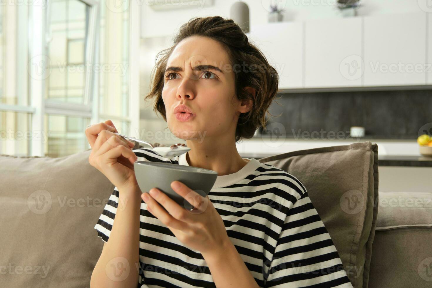 retrato do menina comendo tigela do cereais e assistindo filme em televisão, olhando às tela com sério face, tendo café da manhã dentro vivo quarto foto