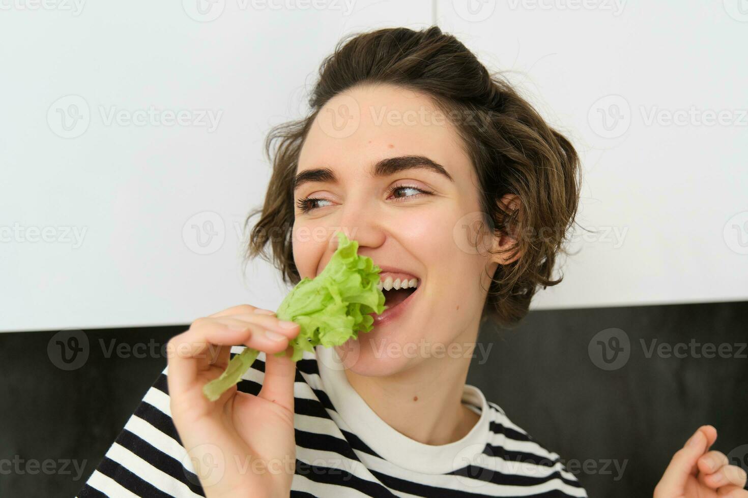 retrato do despreocupado vegetariano garota, comendo vegetais, mordida alface folha com feliz sorridente face, tendo saudável lanche, curtidas vegetais, carrinhos dentro a cozinha foto