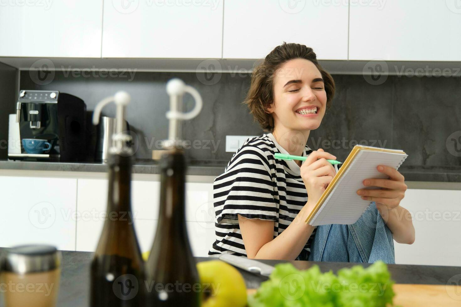 fechar acima do feliz, sorridente jovem mulher escrevendo dentro caderno, criando receita, Lista do refeições para isto semana, sentado perto legumes e Oliva óleo, cozinhando dentro a cozinha foto