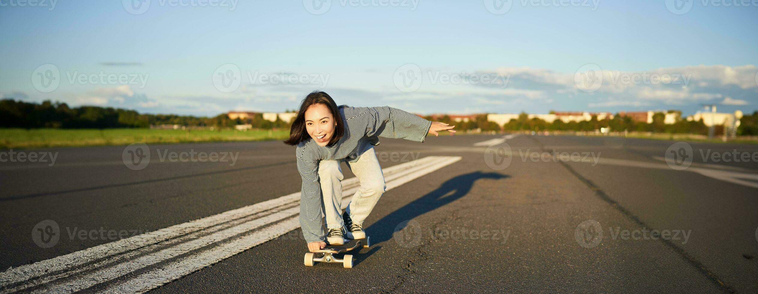 despreocupado skatista menina em dela skate, equitação Longboard em a esvaziar estrada, segurando mãos lateralmente e rindo foto