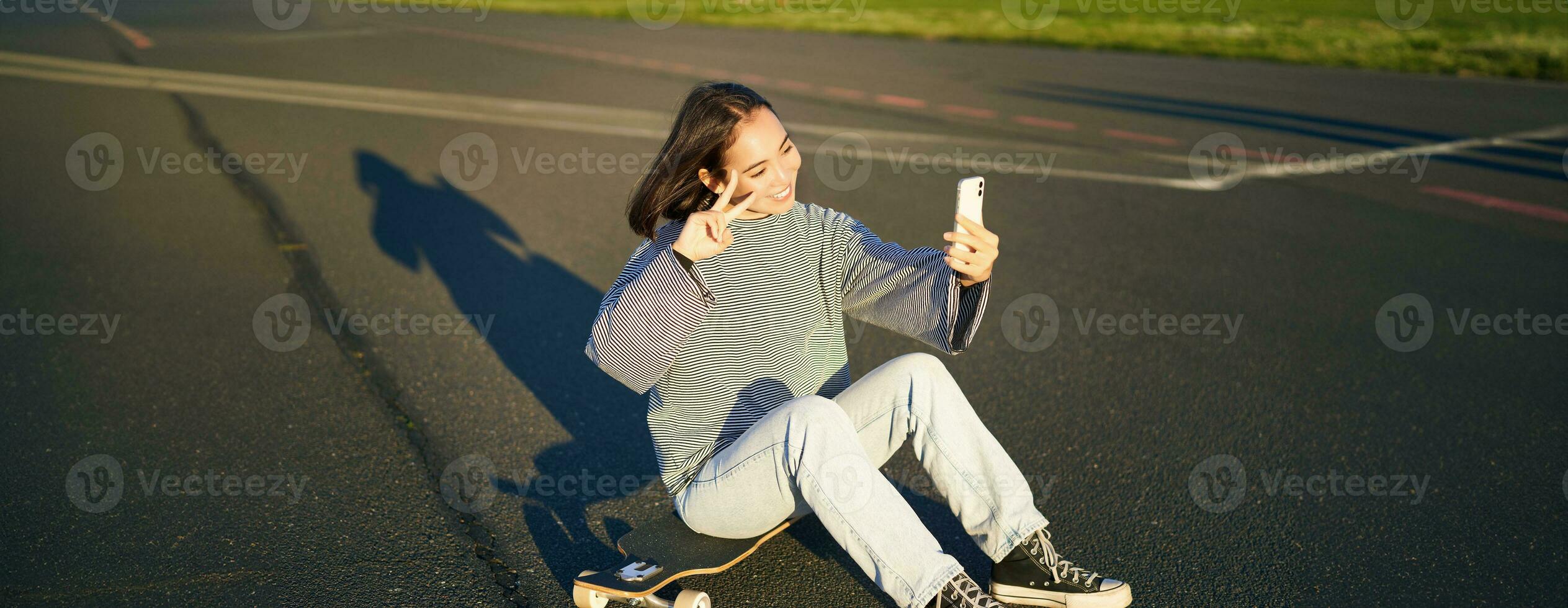 feliz ásia menina senta em skate, leva selfie com Longboard, faz fofa rostos, ensolarado dia ao ar livre foto