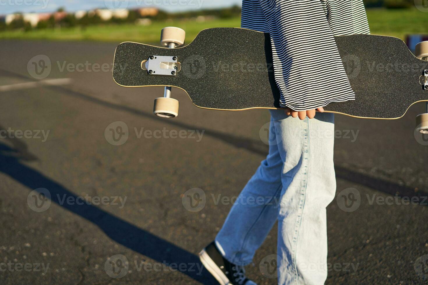 cortada tiro do adolescente skatista garota, mãos segurando Longboard, caminhando com skate em concreto estrada foto