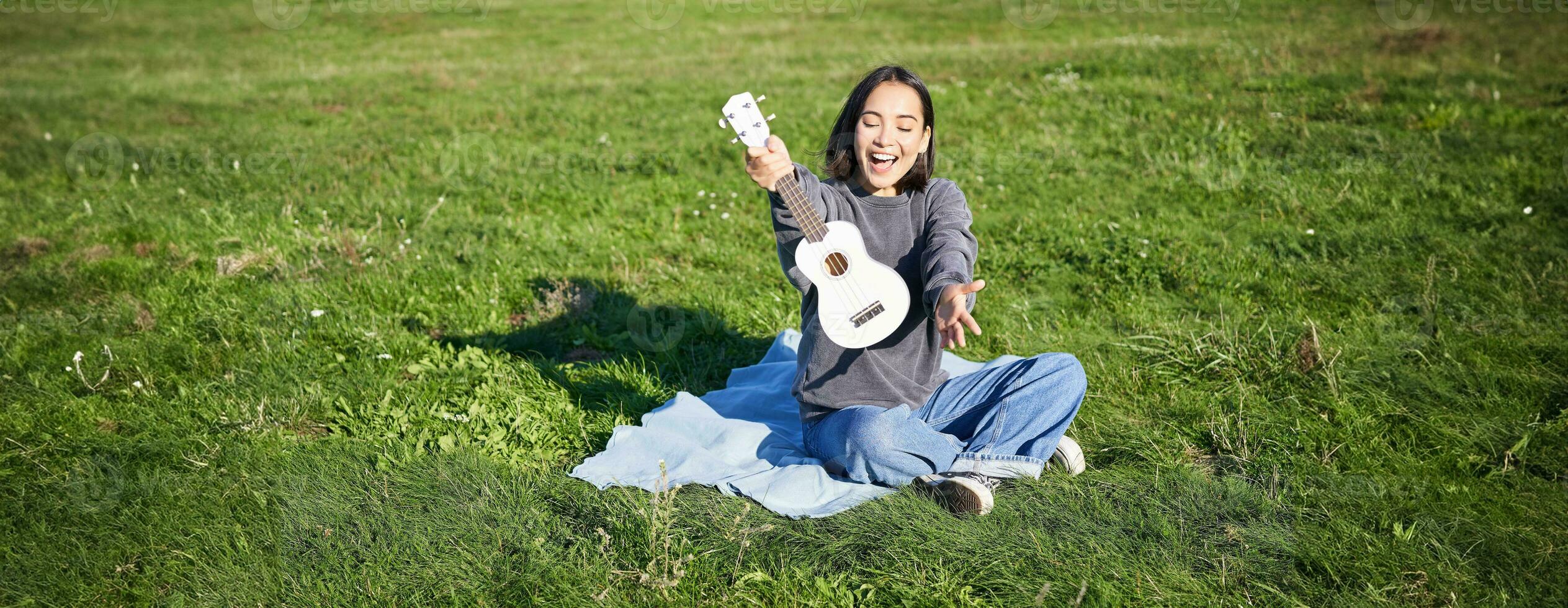 música e instrumentos. retrato do fofa ásia menina mostra dela branco cavaquinho, tocam dentro parque enquanto sentado relaxado em cobertor, desfrutando ensolarado dia foto