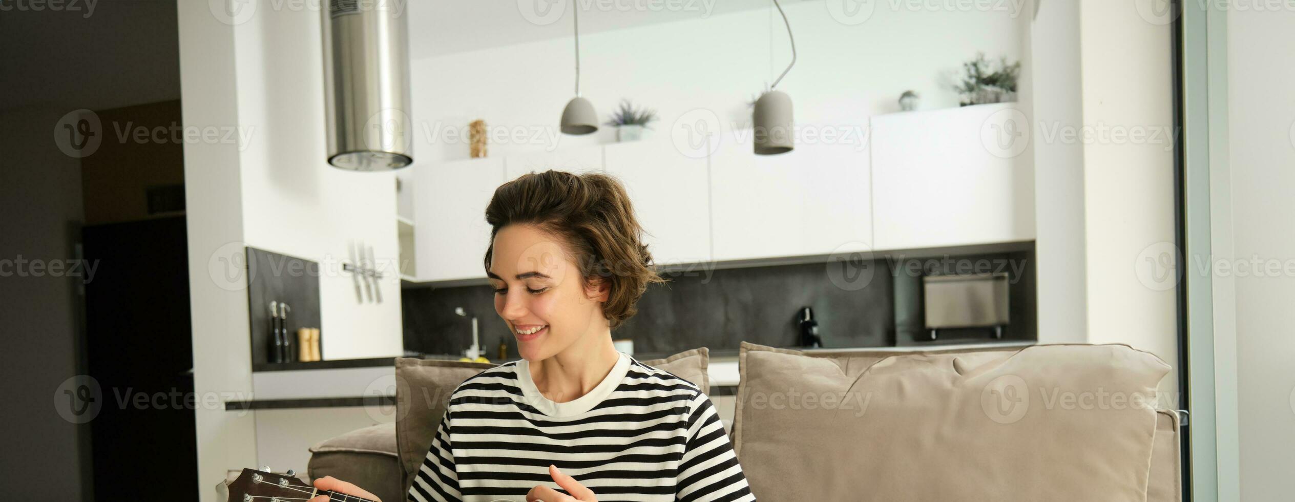 vertical retrato do feliz jovem mulher jogando cavaquinho, aprende Novo musical instrumento, senta em sofá às lar, sorridente com alegria foto