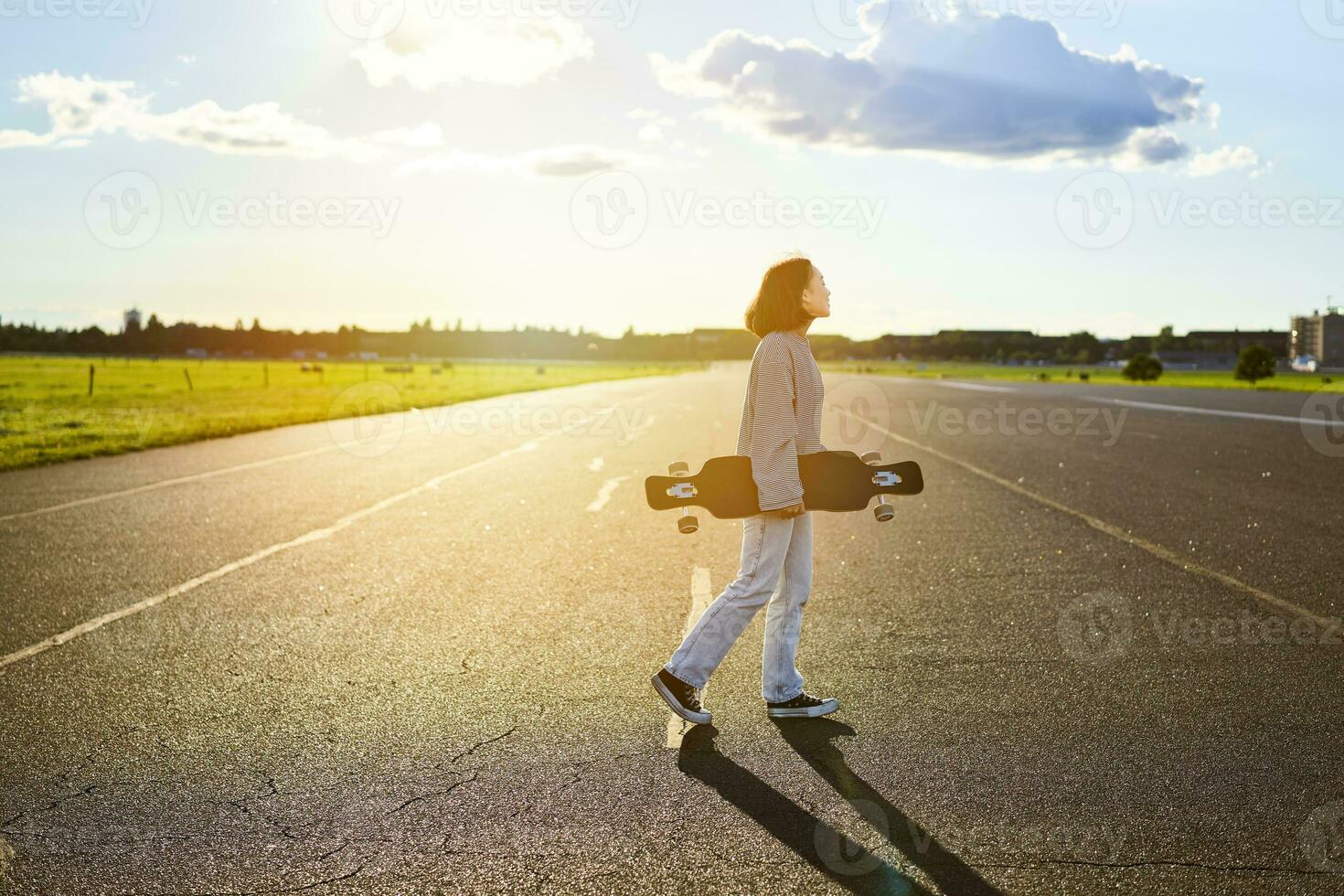 jovem skatista garota, adolescente patinação em cruzador, segurando Longboard e caminhando em concreto esvaziar estrada foto