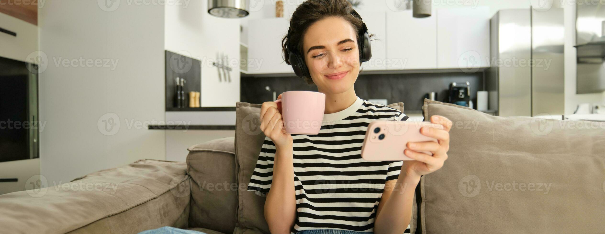 sorridente menina assistindo televisão mostrar em Smartphone, vestindo fones de ouvido, bebendo chá e sentado em sofá foto