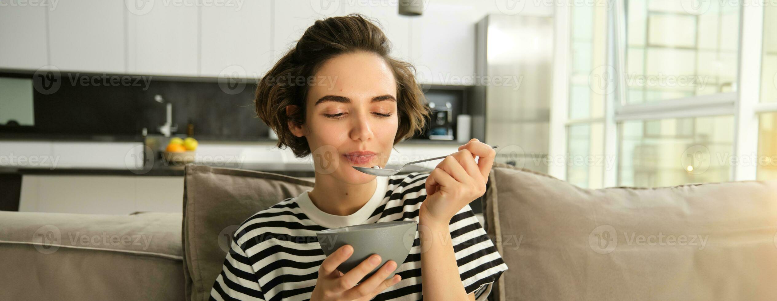 retrato do jovem mulher comendo granola, tigela do cereais com leite, sentado em sofá e tendo dela café da manhã foto