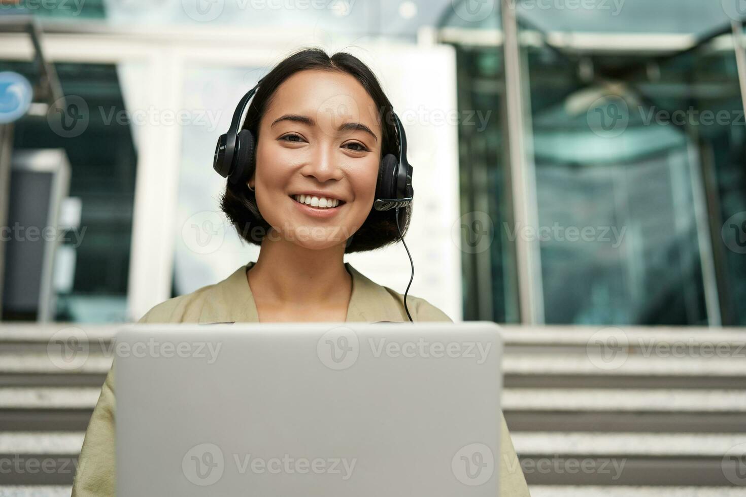 retrato do jovem ásia mulher sentado com computador portátil e fones de ouvido, assistindo vídeo, faz conectados curso em computador, sentado em escadas ao ar livre foto
