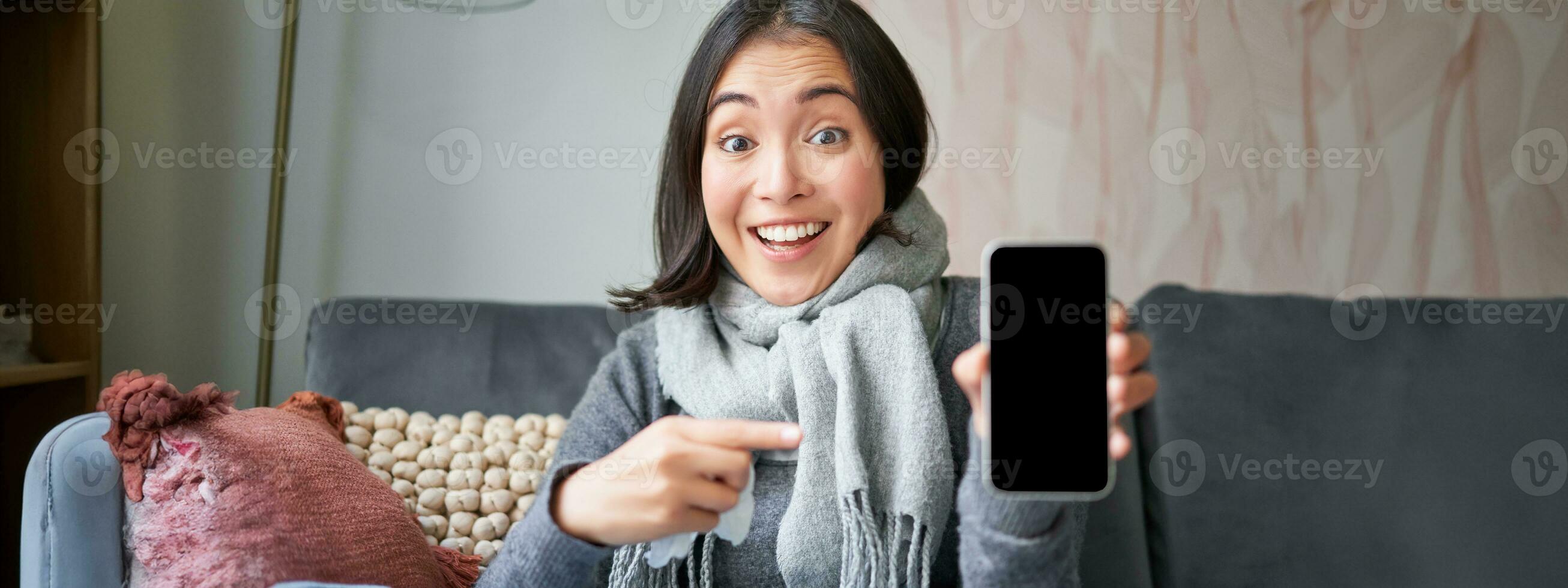 animado jovem mulher apontando dedo às Smartphone, mostrando conectados doutor, médico inscrição ou gp contato em Móvel telefone, ficando às casa doente, pegando frio foto