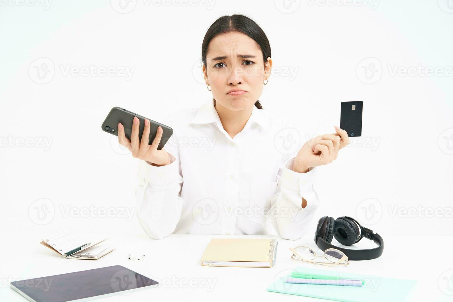 retrato do ásia mulher senta às mesa, detém crédito cartão e Smartphone, parece chateado, desapontado de preços, branco fundo foto