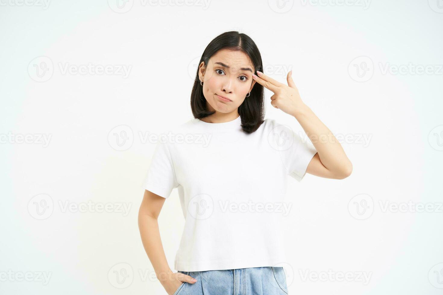 irritado ásia mulher, aluna mostra dedo pistola perto cabeça, parece incomodado e cansado, carrinhos isolado em branco fundo foto