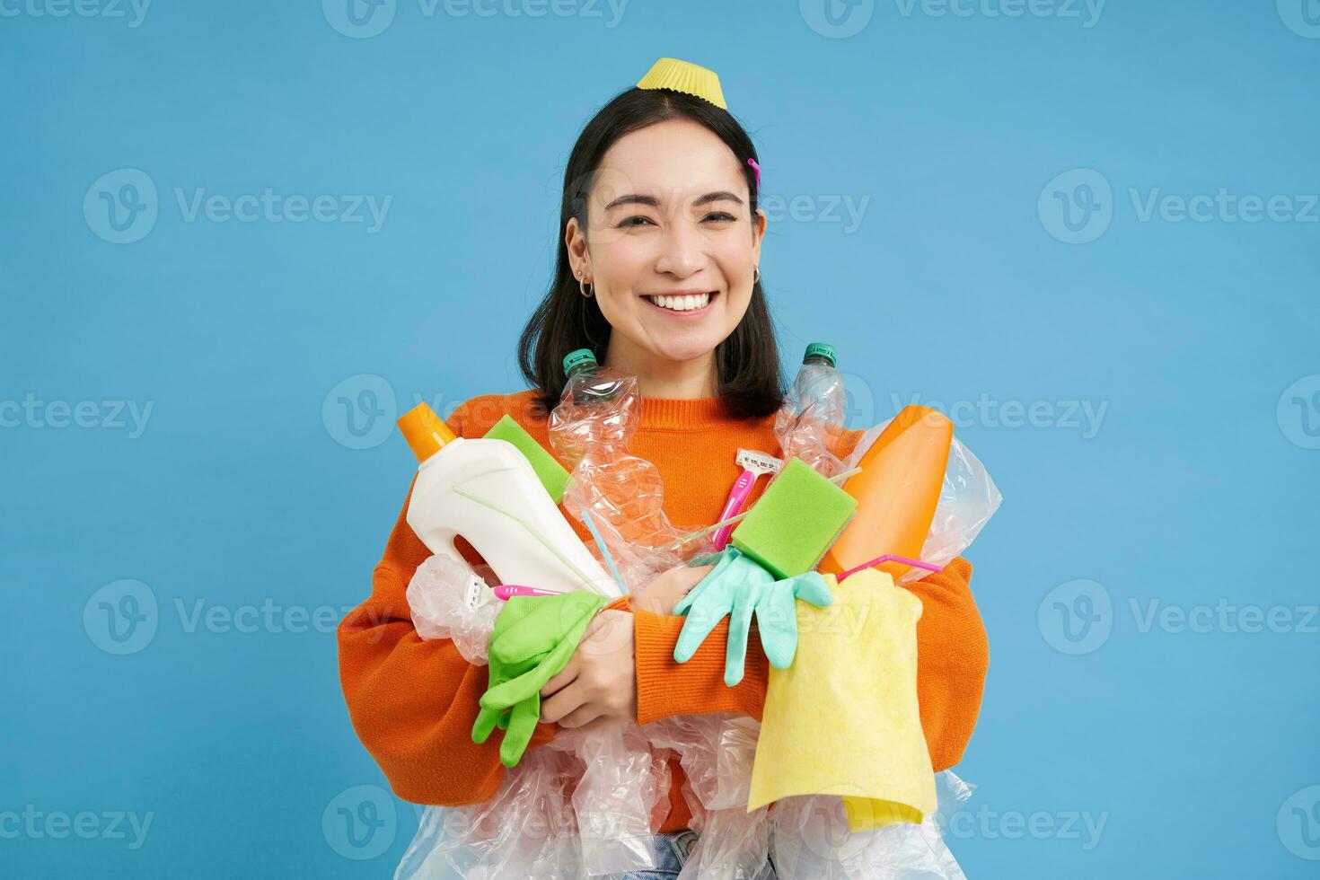 retrato do sorridente coreano mulher, segurando esvaziar plástico garrafas, lixo para reciclando, olhando entusiasmado, Ordenação Fora lixo, azul fundo foto