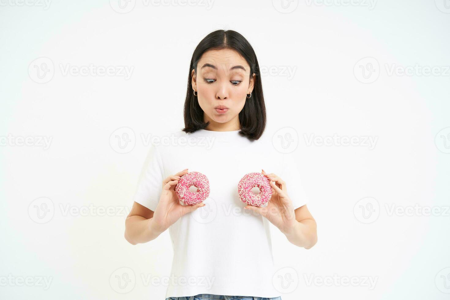 retrato do jovem engraçado mulher, segurando dois Rosa envidraçado rosquinhas sobre peito, parece espantado às sobremesa, branco fundo foto