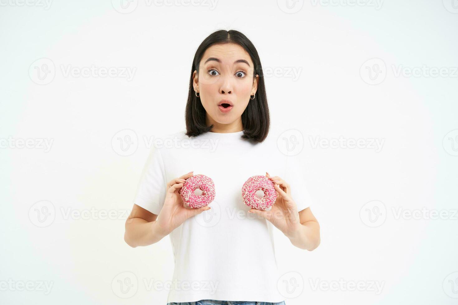 retrato do mulher com surpreso face, segurando dois rosquinhas sobre seio e olhando chocado, branco fundo foto