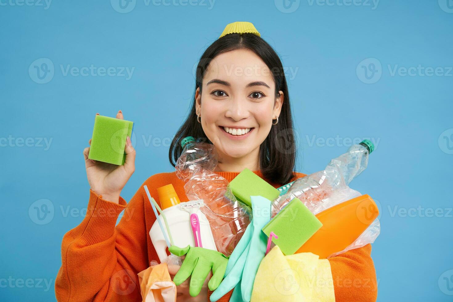 entusiasmado ásia menina mostrando limpeza esponja, segurando esvaziar plástico garrafas, Lixo para reciclando, Ordenação dela família lixo, azul fundo foto