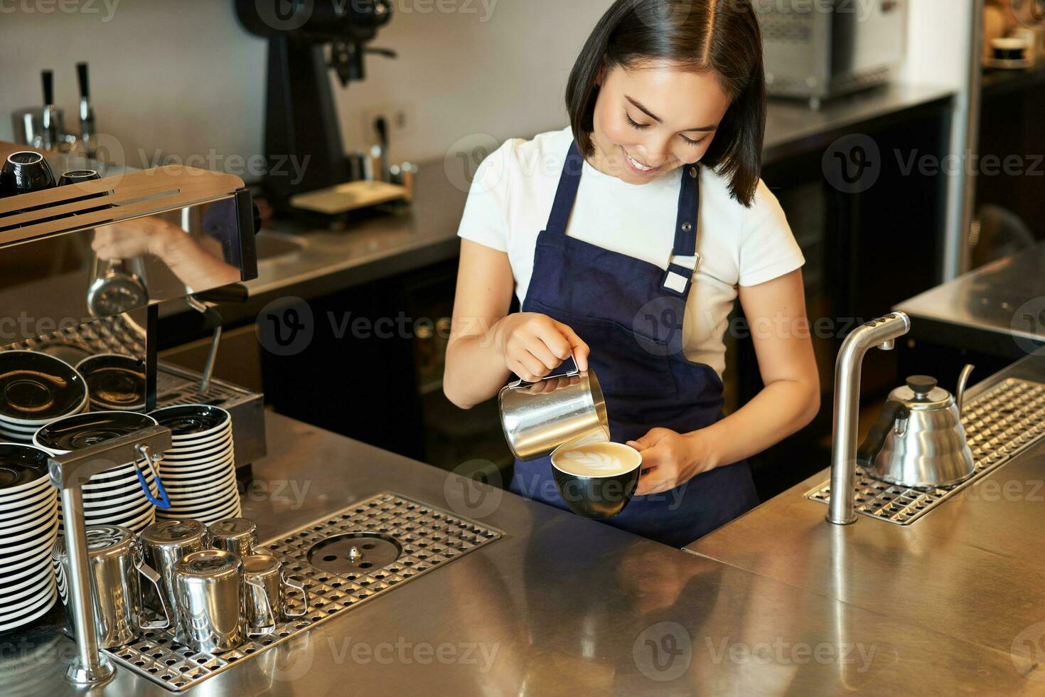 sorridente ásia menina barista, cafeteria funcionários derramando cozido no vapor leite dentro café, preparar cappuccino com café com leite arte, em pé dentro azul avental atrás contador foto