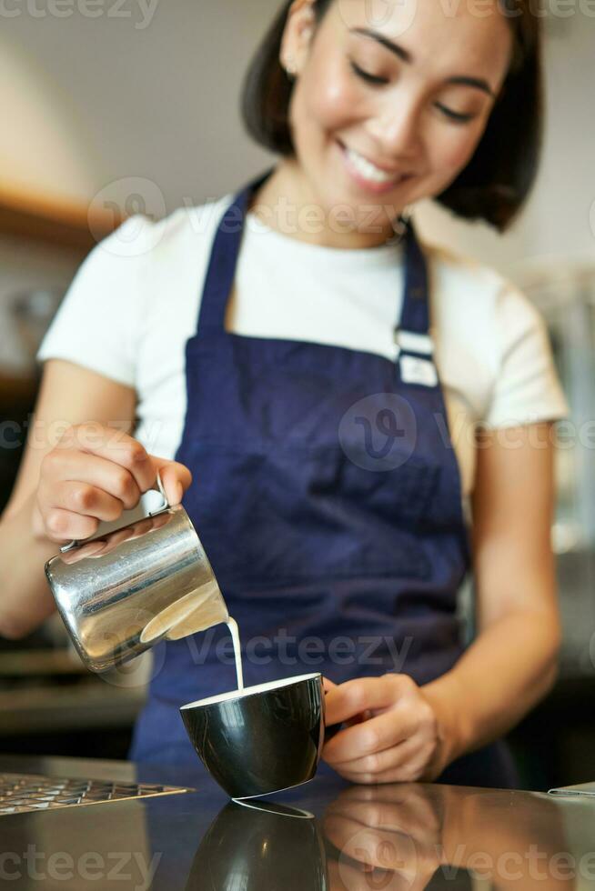 vertical tiro do morena ásia menina barista, vestindo avental, fazer café, preparar ordem dentro cafeteria, derramando leite dentro copo para cappuccino café com leite arte foto