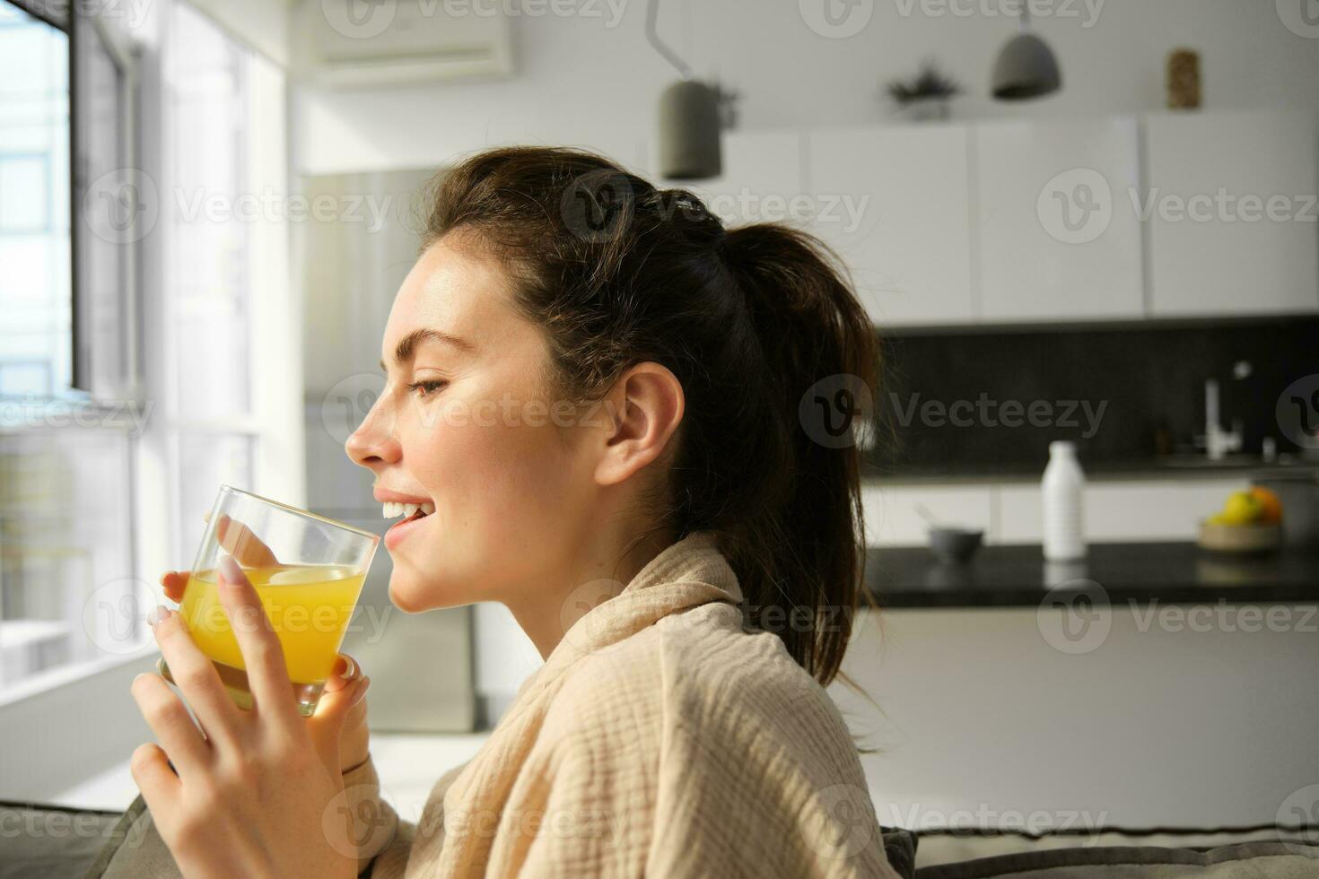 fechar acima retrato do feliz jovem mulher, goza relaxante dia às lar, bebidas vidro do laranja suco e sorrisos, parece lado de fora janela foto