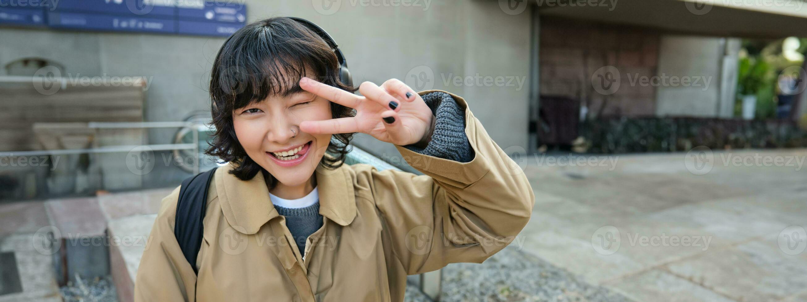 cabeça retrato do coreano menina em pé em rua, mostrando Paz sinal, vestindo fones de ouvido, ouvindo música e sorridente foto