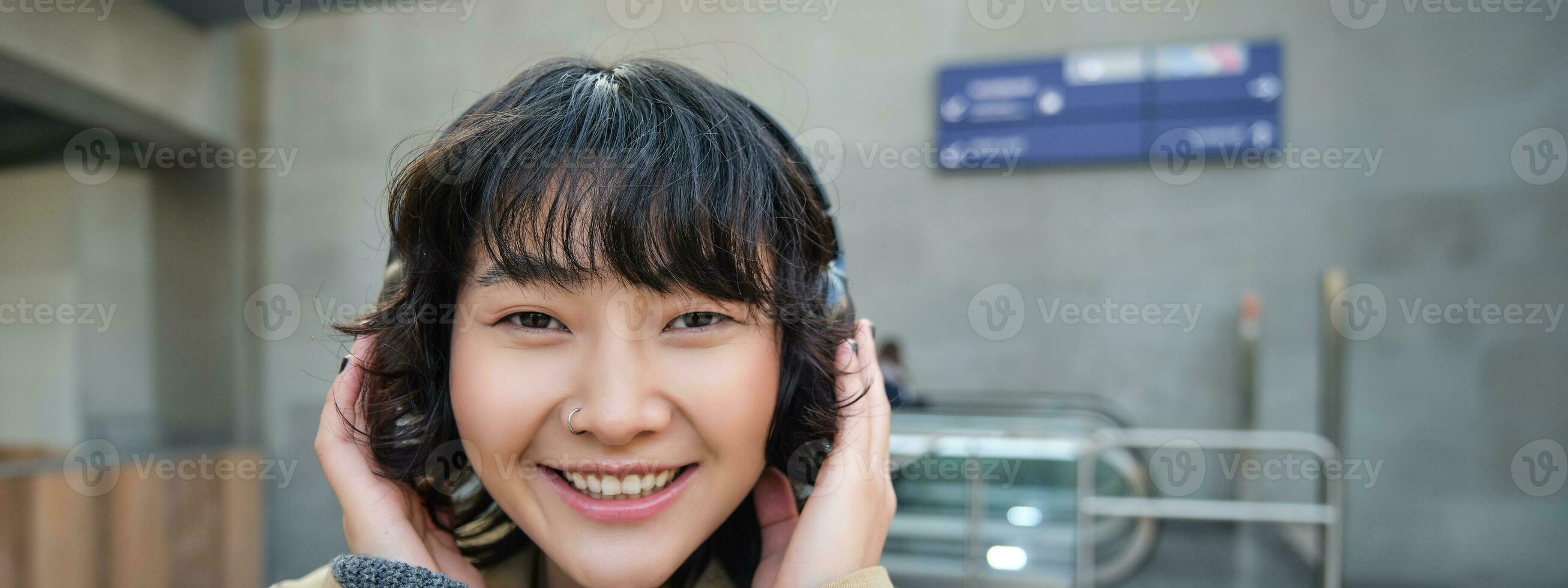 fechar acima retrato do à moda coreano menina em pé em rua, ouvindo música dentro fones de ouvido e sorridente, posando dentro cidade Centro foto