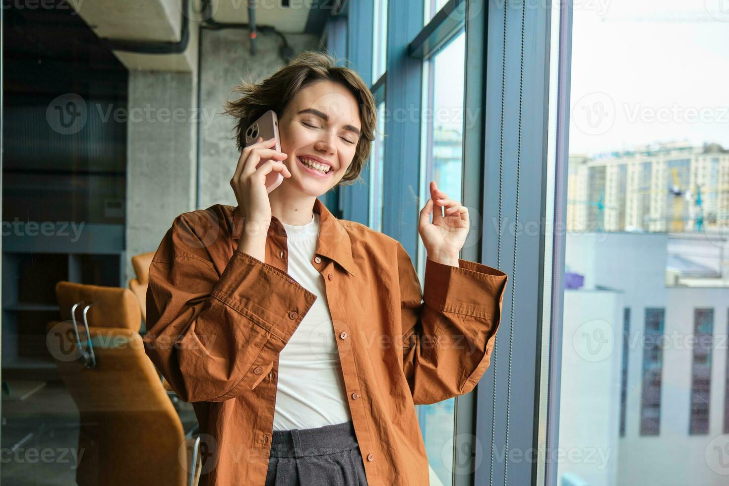 retrato do trabalhando mulher, social meios de comunicação Gerente dentro escritório, falando em Móvel telefone, tendo conversação em Telefone, em pé Próximo para a janela foto