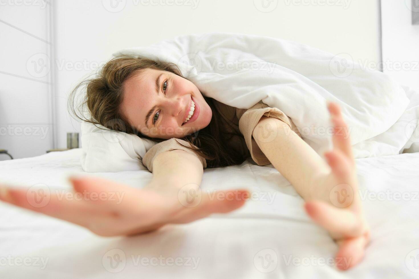 alegre menina alongamento dela braços dentro cama, acordando acima a partir de Boa dormir ou sesta, desfrutando a manhã foto